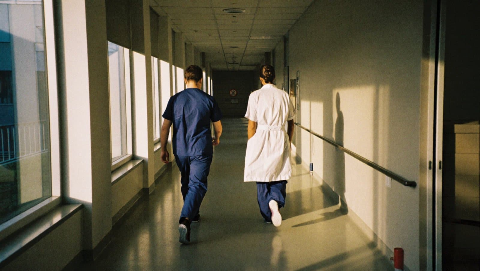 Two clinicians walk away down a sunlit hospital corridor in the early morning.